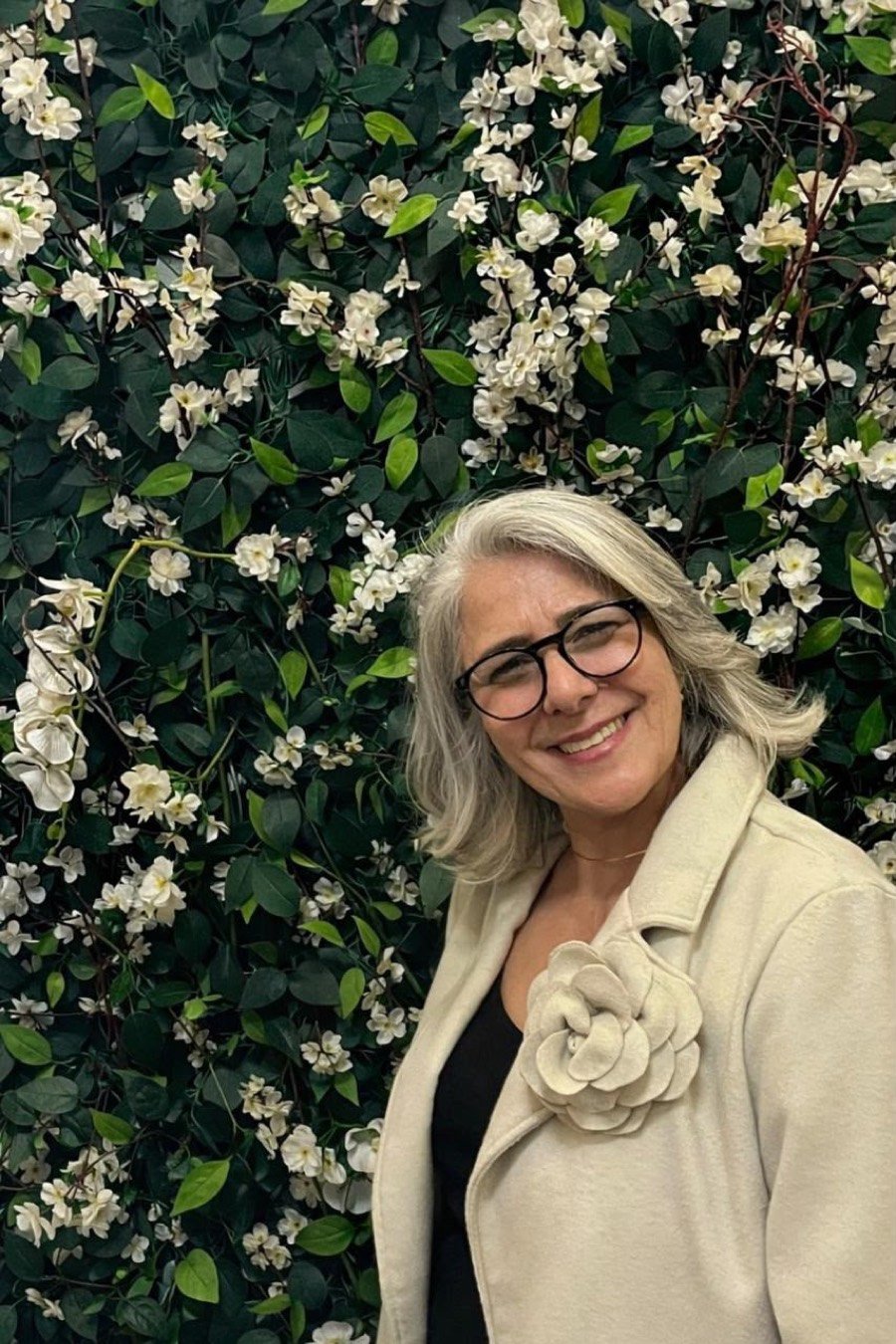 Lú Peixoto sorrindo em frente a um jardim de flores brancas